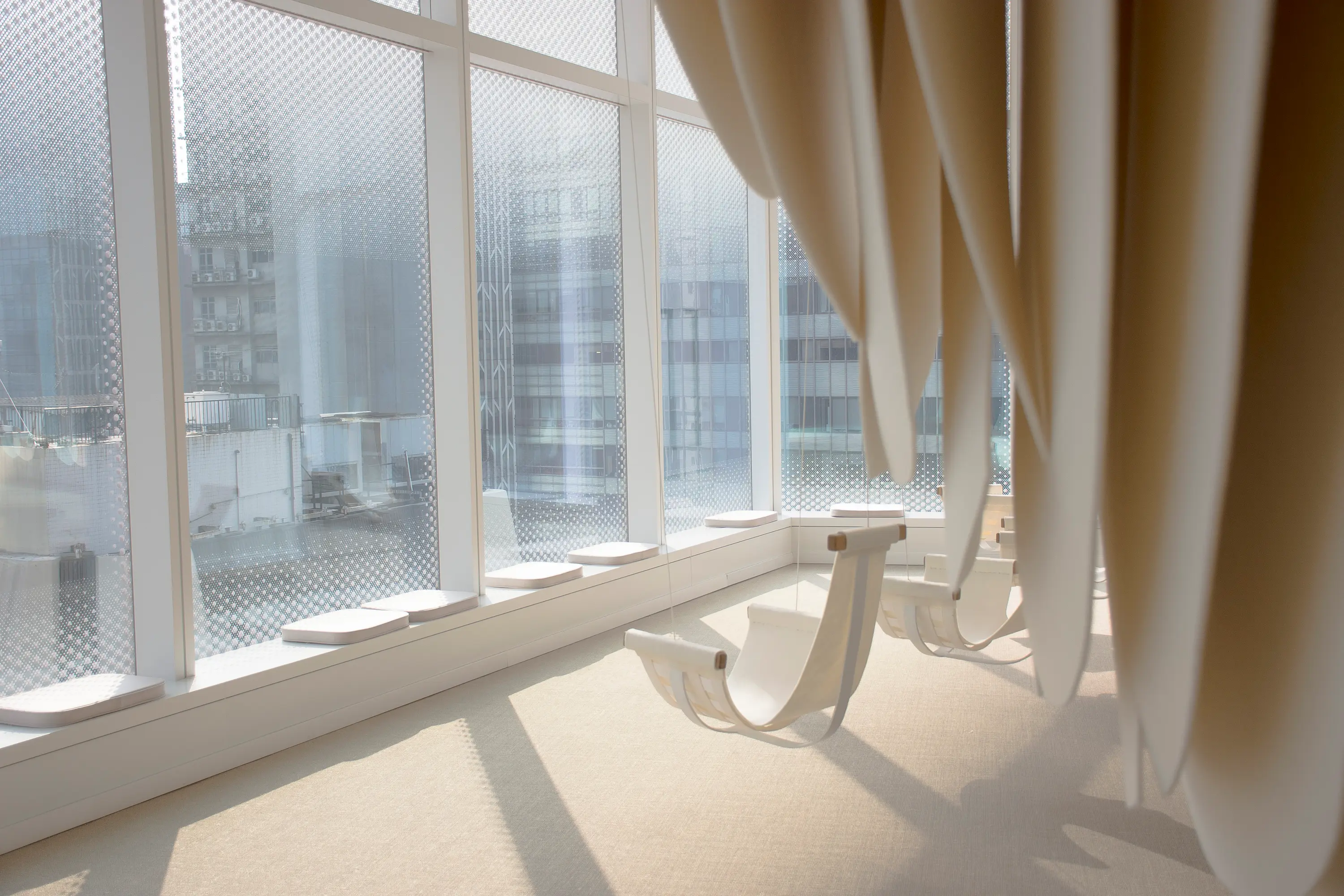 A light-filled installation space with sculptural seating and hanging white forms beside large windows overlooking the city.